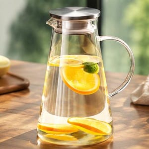 Peut inclure: Une carafe en verre transparent avec un corps conique, une anse en verre courbée et un couvercle en métal argenté. La carafe est remplie d'eau et contient plusieurs tranches d'agrumes orange vif et un petit quartier de citron vert flottant.