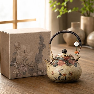Może przedstawiać: Ceramiczny czajniczek w kremowe kropki, ozdobiony malowanym kotem i motywami kwiatowymi. Posiada ciemny, owinięty uchwyt z małą zawieszką oraz pasującą pokrywkę. Obok znajduje się kartonowe pudełko ozdobione ilustracjami kwiatów.