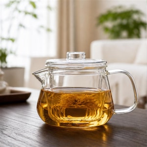 May include: A clear glass teapot with a matching lid and a side handle. The teapot contains a golden-coloured liquid with a single submerged yellow flower blossom visible through the transparent walls of the vessel, showcasing its simple design.