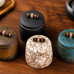 May include: Four cylindrical ceramic canisters with lids, each featuring a small dark brown wooden handle tied with twine. The canisters come in solid black, mottled brown, white with brown speckles, and teal with dark brown textured patterns.