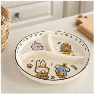 May include: A round, cream-colored ceramic divided plate featuring three sections. The rim is decorated with small yellow stars and swirls. The plate surface displays colorful illustrations of a pig, a bear, a rabbit, and an elephant.