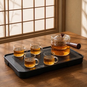 May include: A clear glass teapot with a dark wooden side handle and lid knob, accompanied by three matching glass teacups filled with amber tea. The set is arranged on a rectangular black slatted tea tray with a drainage design for serving.
