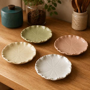 May include: A set of four small ceramic plates in pale green, soft pink, cream, and white. Each plate features a circular shape with a scalloped, ruffled edge and a central embossed pattern resembling the veins of a leaf or a flower petal.