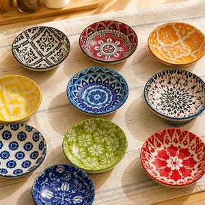 Può includere: Una collezione di dieci piccole ciotole rotonde in ceramica disposte in una griglia. Ogni ciotola presenta un motivo unico e colorato, inclusi disegni geometrici, floreali e ispirati ai mandala nei toni del rosso, blu, giallo, verde, arancione, nero e bianco.