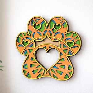 May include: A layered wooden paw print wall art piece. The design features a heart-shaped cutout and smaller hearts within the paw pads. The artwork uses orange, green, blue, and yellow colors, creating a vibrant and decorative piece.