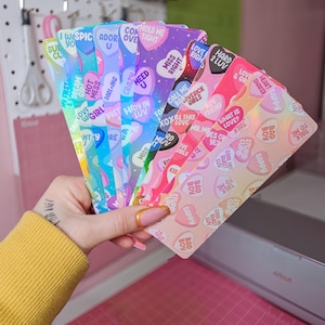 May include: A hand holds a fan of colorful, heart-shaped cards with various phrases like "Hold Me Tight" and "Love Right." The cards have a holographic sheen and are in shades of pink, blue, and purple. The background includes a pegboard and a pink cutting mat.