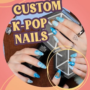 May include: Custom K-Pop nails in light blue with cloud and flower designs. One nail features a teddy bear. The nails are displayed against a brown background with a pink and orange gradient.