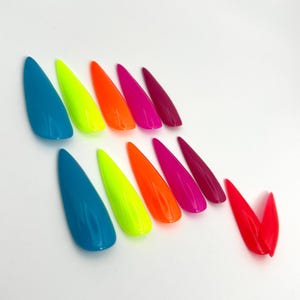 May include: A collection of long, pointed artificial nails in various vibrant colors. The colors include teal, neon yellow, orange, hot pink, and deep magenta. The nails are arranged on a white surface.