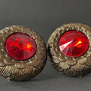 May include: A pair of vintage-style earrings. Each earring features a large, faceted red gemstone set in a round, bronze-colored metal frame with a detailed, textured design. The earrings are displayed against a dark background.