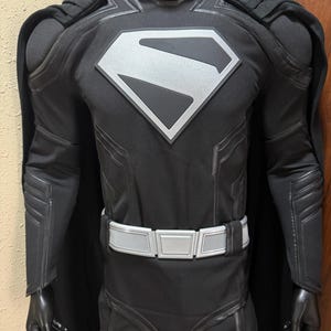 Può includere: Costume da supereroe nero con un emblema argentato sul petto. Il costume presenta un mantello nero, una cintura argentata e pannelli dettagliati su braccia e gambe. L'abito è esposto su un manichino.