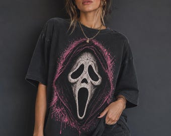 Op de afbeelding: Een antracietgrijs T-shirt met korte mouwen en een grote, centrale afbeelding van een wit, spookachtig masker met een zwarte capuchon, omringd door een felroze spettereffect. Het T-shirt heeft een ruime pasvorm en een vervaagde afwerking.