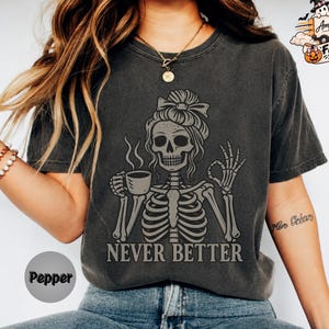 May include: A charcoal grey short-sleeve t-shirt featuring a graphic of a skeleton with a messy bun hairstyle, holding a steaming cup of coffee and making an okay hand gesture. The text "NEVER BETTER" is printed in grey below the skeleton.