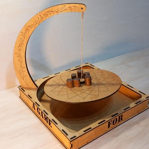 Op de afbeelding: Houten spel met een gebogen boog en een cirkelvormig platform. Het platform heeft een geometrisch ontwerp en bevat kleine houten cilinders. De basis van het spel heeft de inscriptie "A GAME FOR". Het spel is gemaakt van lichtbruin hout.
