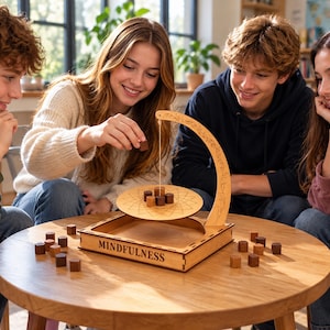 Könnte beinhalten: Ein hölzernes Balancierspiel mit einem quadratischen Sockel, auf dessen Vorderseite das Wort MINDFULNESS eingraviert ist, einem geschwungenen Arm und einer hängenden kreisförmigen Plattform. Kleine braune Holzwürfel liegen auf der Plattform.