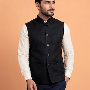 Può includere: Un gilet nero con collo alla coreana e chiusura con bottoni. Il gilet ha una tasca sul petto e viene indossato sopra una camicia bianca a maniche lunghe. Il modello indossa pantaloni blu navy.