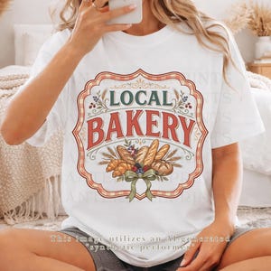 Può includere: Una t-shirt bianca a maniche corte con una grafica centrale che riporta il testo LOCAL BAKERY in rosso e verde. Il design include illustrazioni di vari tipi di pane, spighe di grano e bacche, incorniciate da un bordo decorativo rosso e bianco.