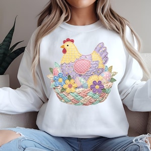 May include: White sweatshirt with an Easter-themed design. The graphic shows a patchwork hen in a basket with decorated eggs and flowers. The hen has yellow, purple, and pink details, and the basket has a woven look. A seasonal, springtime garment.