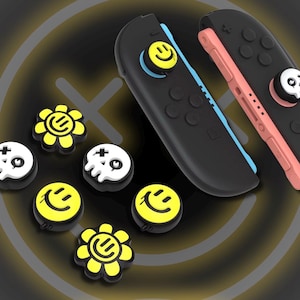 Puede incluir: Mandos de juego negros con agarres para el pulgar amarillos con forma de calavera y emoticono. También se ven agarres para el pulgar adicionales con forma de flor y emoticono amarillos. Los mandos tienen detalles en azul y rosa.