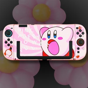 May include: A pink Nintendo Switch controller with a cartoon Kirby design. The controller has black joysticks and buttons, with the text "PINK PUFF ROW" on the left side. The background features pink flower-shaped decorations.
