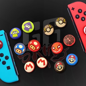 Puede incluir: Una colección de coloridas tapas de joystick con varios diseños, incluyendo un hongo rojo, un hongo azul, un logo de Spider-Man y una cara de Mario. Las tapas se muestran con un mando de juego rojo y azul.