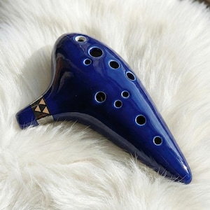 May include: A glossy, cobalt blue ocarina with multiple finger holes. The ceramic wind instrument has a rounded shape with a mouthpiece and a decorative triangle pattern in black, gold, and silver.
