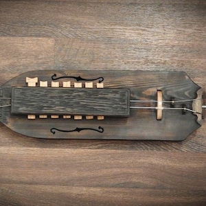 Puede incluir: Un instrumento musical de cuerda, una zanfona de madera con acabado oscuro. Cuenta con una manivela, cuerdas y teclas. El cuerpo es de color marrón oscuro con tallas decorativas y una caja de resonancia rectangular.