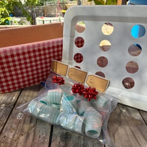 May include: A clear plastic bag containing multiple small, light blue cylindrical plastic caps, decorated with three small red bows and three blank tan gift tags labeled To and From, positioned in front of a white plastic grid board.