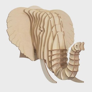 May include: A light-coloured, wooden elephant head sculpture. The sculpture is made from layered wooden pieces, creating a 3D effect. The elephant has large ears, tusks, and a detailed trunk. The artwork is designed to be mounted on a wall.