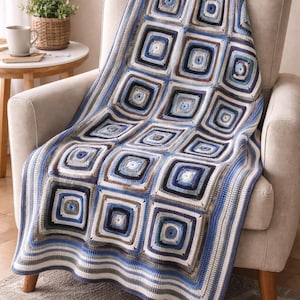 May include: A blue, white, and brown crocheted blanket with a geometric square pattern draped over a beige armchair. The blanket features a repeating design of squares within squares, creating a textured appearance. The color palette includes shades of blue, white, and brown.