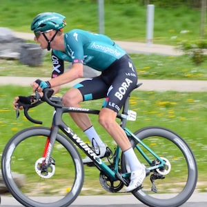 May include: A cyclist in teal and black cycling gear, including a helmet and sunglasses, rides a Specialized S-Works road bike. The bike has black wheels with red accents and the cyclist is in motion on a green grassy area.