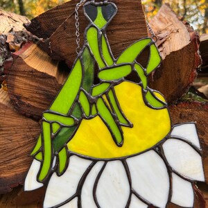 May include: A stained glass praying mantis with a green body, black eyes, and yellow and white flower. The insect is perched on a yellow center and white petals. A silver chain is attached for hanging. The artwork is set against a backdrop of wood.