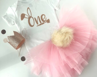 Puede incluir: Un tutú de tul rosa con un pompón de piel blanca, un body blanco con el texto "One" en purpurina dorada y orejas de conejo, y una corona de purpurina dorada.