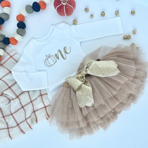 May include: A white long-sleeved onesie with a gold pumpkin and the word "one" printed on the front. A brown tulle skirt with a gold glitter bow is also pictured.