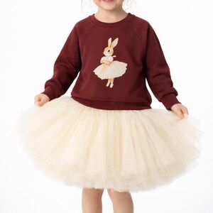 May include: A burgundy sweatshirt featuring a cartoon bunny in a ballerina outfit. The bunny is wearing a white tutu. The sweatshirt is paired with a cream-colored tulle skirt, creating a playful and whimsical ensemble for children.