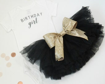 May include: A white bodysuit with the text "BIRTHDAY girl" in black lettering, paired with a black tulle tutu with a gold glitter bow.