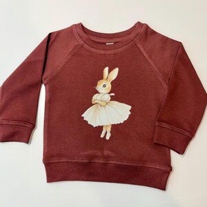 May include: A burgundy-colored children's sweatshirt with raglan sleeves. The front features a whimsical illustration of a bunny in a white tutu and ballet shoes. The bunny has its arms crossed and is wearing a white dress.