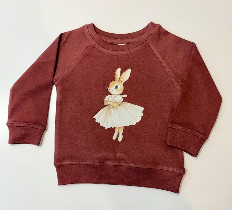 May include: A burgundy-colored children's sweatshirt with raglan sleeves. The front features a whimsical illustration of a bunny in a white tutu and ballet shoes. The bunny has its arms crossed and is wearing a white dress.