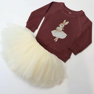 May include: A burgundy long-sleeved sweatshirt and a cream-colored tutu skirt. The sweatshirt features a graphic of a bunny wearing a ballet dress. The tutu is made of layers of soft tulle. The outfit is designed for children.