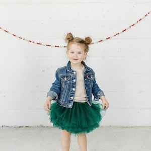 May include: A young child wearing a denim jacket, a beige ribbed top, and a layered emerald green tulle skirt. The child is also wearing white socks. A decorative garland hangs in the background.