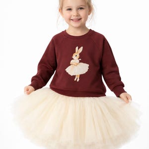 May include: A young girl wearing a burgundy sweatshirt with a bunny ballerina graphic and a cream-colored tulle skirt. The bunny is wearing a tutu. The girl is smiling and looking at the camera.