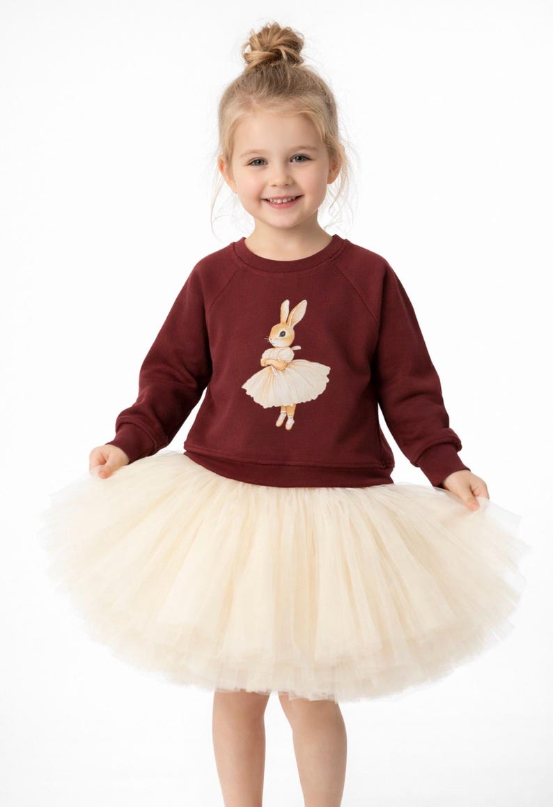 May include: A young girl wearing a burgundy sweatshirt with a bunny ballerina graphic and a cream-colored tulle skirt. The bunny is wearing a tutu. The girl is smiling and looking at the camera.