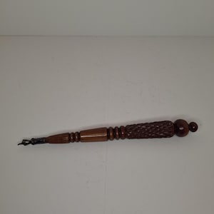 Może przedstawiać: Zabytkowe drewniane pióro maczane z ciemnobrązowym wykończeniem. Pióro ma szczegółowy, rzeźbiony wzór na uchwycie i metalową stalówkę. Pióro ma około 25 cm długości.