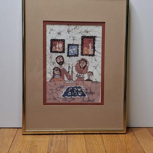 May include: Framed artwork depicting a family gathered around a table, with candles and a Hebrew inscription. The artwork features a gold frame, a tan mat, and a batik-style image with a family and three smaller framed images.