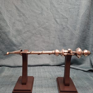 Può includere: Una bacchetta in metallo argentato con un design a spirale e accenti sferici, esposta su due supporti in legno marrone. La bacchetta ha una punta e sfere decorative all'estremità. Lo sfondo è un tessuto strutturato color verde acqua.