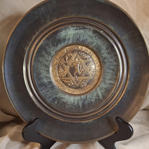 May include: A decorative bronze plate with a central gold medallion featuring a Star of David and Hebrew text. The plate has a dark green and bronze patina, and is displayed on a black stand.