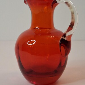 May include: A small, red glass pitcher with a clear handle. The pitcher has a rounded body and a flared rim. The handle is attached to the top and bottom of the pitcher. The glass is smooth and glossy.