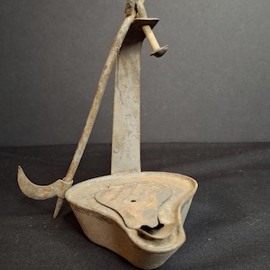 May include: A weathered, gray metal oil lamp featuring a triangular reservoir with a hinged lid and a central wick opening. An upright metal support arm is attached to the back, holding a long, slender metal rod with a curved hook at the end.