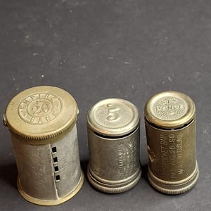 Könnte beinhalten: Drei zylindrische Taschenspardosen aus Metall mit messingfarbenen Kappen und Sockeln. Die linke Spardose ist mit Empire Bank 20 beschriftet. Die mittlere zeigt eine 5 auf der Oberseite. Die rechte ist mit Six Pence und Pocket Bank beschriftet.