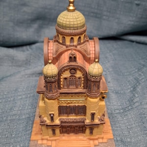 May include: A detailed miniature replica of a building with a domed roof and multiple turrets. The model features a yellow facade with brown and gold accents, and a light green dome. The architectural model sits on a rectangular base.