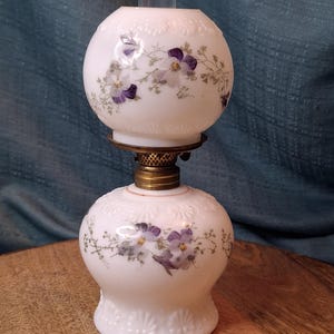 Op de afbeelding: Een vintage witte olielamp met een ronde bol en voet, versierd met paarse bloemmotieven. De lamp heeft een messing fitting en een heldere glazen schoorsteen. De voet heeft een decoratief reliëfpatroon.